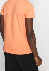 Persona de piel oscura vistiendo una camiseta de manga corta color durazno y pantalones negros, de pie contra un fondo claro y liso, con la espalda visible.