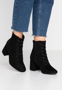 Stivali ankle in suede neri con punta appuntita, chiusura a lacci sul davanti e zip laterale. Tacco chunky con superficie testurizzata.