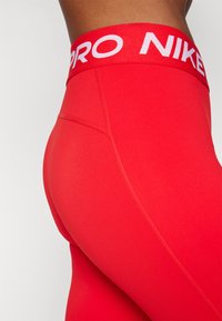 Czerwone legginsy Nike Pro z wysokim pasem i białym logo, wykonane z gładkiego, elastycznego materiału, zaprojektowane dla komfortu i swobody ruchów.