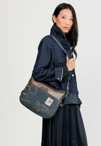 Borsa a spalla blu scuro con un accento marrone testurizzato, presenta stampe decorative e accessori metallici. Manico regolabile e tasche funzionali.