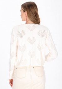 Häkelstrickpullover in Creme mit V-förmigem Muster und gerippten Bündchen, kombiniert mit einem hellbeigen Rock mit Gesäßtaschen.