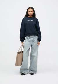 Σκούρο μπλε φούτερ με κεντημένο το "Tommy Hilfiger", συνδυασμένο με ανοιχτό μπλε φαρδύ τζιν. Μπεζ τσάντα κρατιέται στο δεξί χέρι.