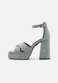 Kang sandalsi denimu sa cross-strap dizajnom, blok petom, kaišem oko gležnja i svijetloplavom bojom, sa suptilnom ispucanom teksturom.