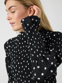 Μαύρο τοπ με λευκές πουά, υφή με σούρες και  φουσκωτά μανίκια. Διαθέτει μαζεμένα μανσέτα και σχέδιο με ψηλό λαιμό.