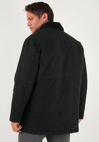 Černý kabát z vlněné směsi s vysokým límcem, dlouhými rukávy a rovný střihem. Má šité detaily na zádech a ramenech.