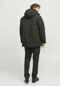 Chaqueta aislante de color verde oscuro con capucha, con un corte recto y tejido texturizado. Llevada sobre pantalones y zapatos negros.