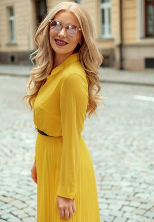 Žluté šaty s dlouhým rukávem a skládáním na sukni, stažené v pase černým opaskem; přehnané růžové sluneční brýle; světlé, vlnité vlasy.