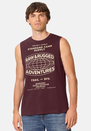 Ung mand med kort brunt hår iført en ærmeløs vinrød skjorte med teksten "Raw & Rugged Adventures" og blå jeans, stående mod hvid baggrund.