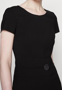 Woman wearing a fitted black dress with short sleeves and a small metallic round buckle detail at the waist.