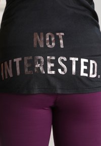 Zwarte t-shirt met vet, metallic roségouden tekst op de achterkant die "NIET GEÏNTERESSEERD" zegt, gecombineerd met strakke paarse leggings.