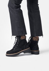 Bottines en suède noir avec un accent en imprimé léopard texturé, lacets à l'avant et semelle en caoutchouc robuste. Jeans noirs avec ourlet déchiré visibles.