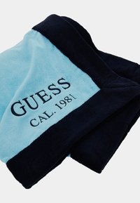 Lyseblå og marineblå plyss tæppe med broderi af "GUESS" og "CAL.1981". Blød tekstur med kontrasterende kanter.