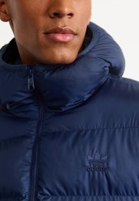 Mere puffer-jakk, millel on kõrge kaelus ja lukuga sulgumine. Õmmeldud Adidas logo rinnal. Sile, läikiv tekstuur koos tikandiga.