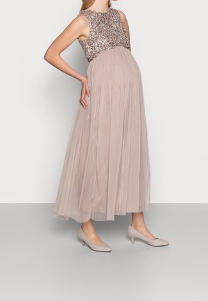 Kobieta w ciąży w sukni bez rękawów z cekinową górą, zwiewną plisowaną spódnicą i beżowymi butami na niskim obcasie, stojąca na jednolitym tle.