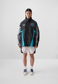 Black and gray zip-up jacket with turquoise accents, signed white shorts, and blue/white shoes. Model stands against a plain background.