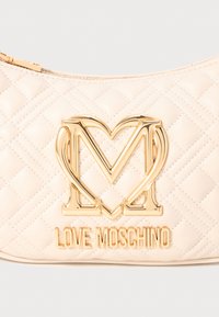 Geantă din piele ecologică bej, matlasată, cu un mare logo în formă de inimă de culoare aurie „M” și numele brandului „LOVE MOSCHINO” embosat în aur dedesubt.