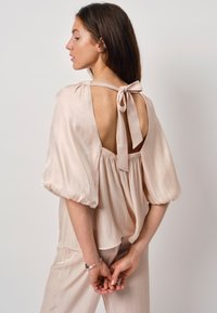 Roze satijnen blouse met een diepe ruguitsnede, gathered details en brede, opbollende mouwen. Met een stoffen lint om de nek gestrikt.