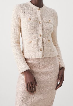 Femme portant une veste crème texturée à boutons avec quatre poches et une jupe assortie texturée sur un fond uni.