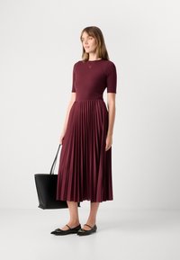 Bordeaux plissert kjole med en tilpasset bodice med korte ermer, teksturert stoff og rund hals. Modell holder en svart totebag.