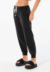 Sort joggingbukser med elastisk talje og justerbar snøre, med sidelommer og tapered manchetter. Bæres med flade sorte sandaler.