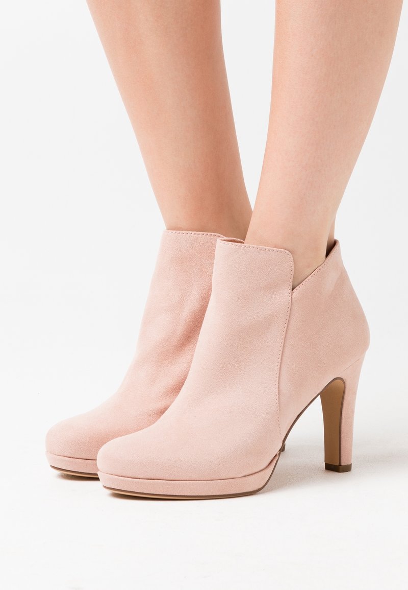 Bottes courtes en daim rose clair à talons hauts portées par une personne se tenant devant un fond blanc.