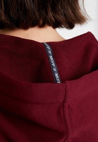 Cheveux bruns mi-longs au niveau des épaules, au-dessus d'un sweat à capuche bordeaux texturé avec une étiquette noire portant l'inscription "MOP" sur la nuque à l'arrière.
