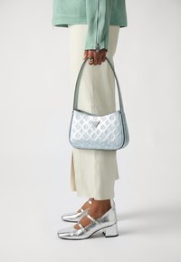 Gaiši zila ādas rokassoma ar iegravētu rakstu, sudraba akcentiem un izliekumu, kuru tur persona, kas ģērbusies piparmētru zaļā krekliņā un krēmkrāsas biksēs.