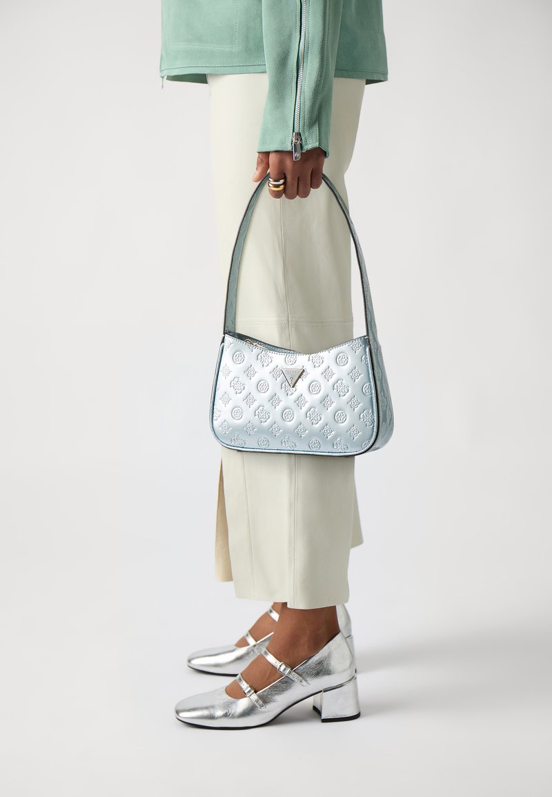 Gaiši zila ādas rokassoma ar iegravētu rakstu, sudraba akcentiem un izliekumu, kuru tur persona, kas ģērbusies piparmētru zaļā krekliņā un krēmkrāsas biksēs.