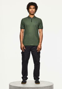 Mężczyzna stojący na okrągłej platformie, ubrany w zieloną koszulkę polo z krótkim rękawem, czarne spodnie cargo i czarne sneakersy z białymi detalami.