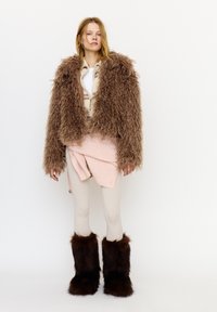 Ung kvinde iført en pjusket brun pelsjakke, cremefarvede leggings, en lyserød sweater bundet om livet og mørkebrune pelse støvler, stående på hvid baggrund.