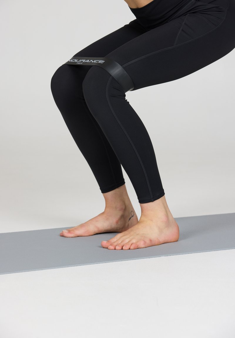 Svarta leggings med sömdetaljer, ett motståndsband placerat över knäna, och barfota på en grå träningsmatta.