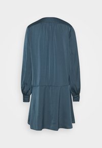 Robe à manches longues bleu sarcelle foncé avec une taille basse et de légers fronces sur le yoke arrière, vue de dos sur un fond uni.