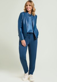 Blauwe leren jas met een hoge kraag, gecombineerd met een satijnen blauwe top en marineblauwe joggingbroek. Witte sneakers maken de outfit compleet.