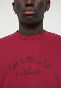 Sweatshirt bordeaux avec texte brodé "ORIGINALS Studio", design à col rond, texture lisse et détails de col côtelé.