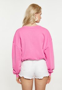 Rózsaszín, oversized pulóver bordázott textúrával. Leeső vállakkal és hosszú ujjakkal rendelkezik. Fehér, foszlott farmer sorttal párosítva.