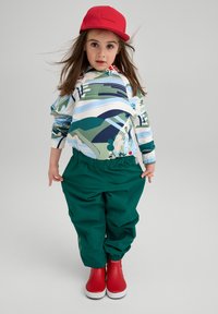 Farverig hættetrøje med abstrakt landskabsdesign, grønne elastiske bukser og røde sko. Indeholder en rød kasket med et logo.