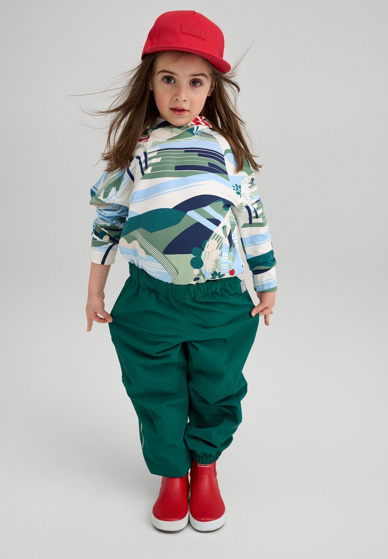 Farverig hættetrøje med abstrakt landskabsdesign, grønne elastiske bukser og røde sko. Indeholder en rød kasket med et logo.