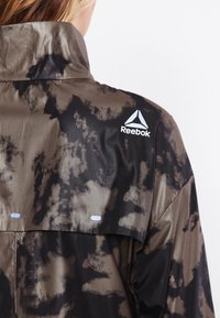 Reebok-jacka med svart och gult mesh-design, hög krage och logotyp. Texturerat tyg med kamouflage-mönster.