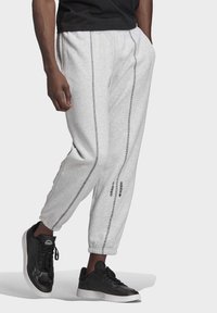 Svetle šedé tepláky s elastickými manžetami, s černými bočními švy a černým logem adidas. Kombinovány s černými teniskami.