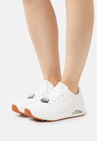 Zapatillas deportivas blancas con una parte superior sintética suave, acentos perforados y suela de goma. Cuentan con cierre de cordones y logotipo de la marca.