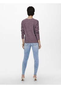 Personne aux cheveux courts et foncés portant un pull en maille violet, un jean skinny bleu clair et des chaussures blanches à talons, debout devant un fond uni.