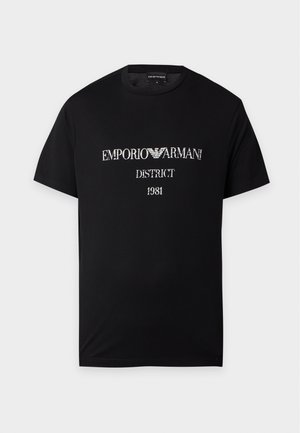 Mężczyzna ubrany na czarno, w czarną koszulkę Emporio Armani, czarne spodnie i czarne trampki, stojący na jednolitym białym tle.