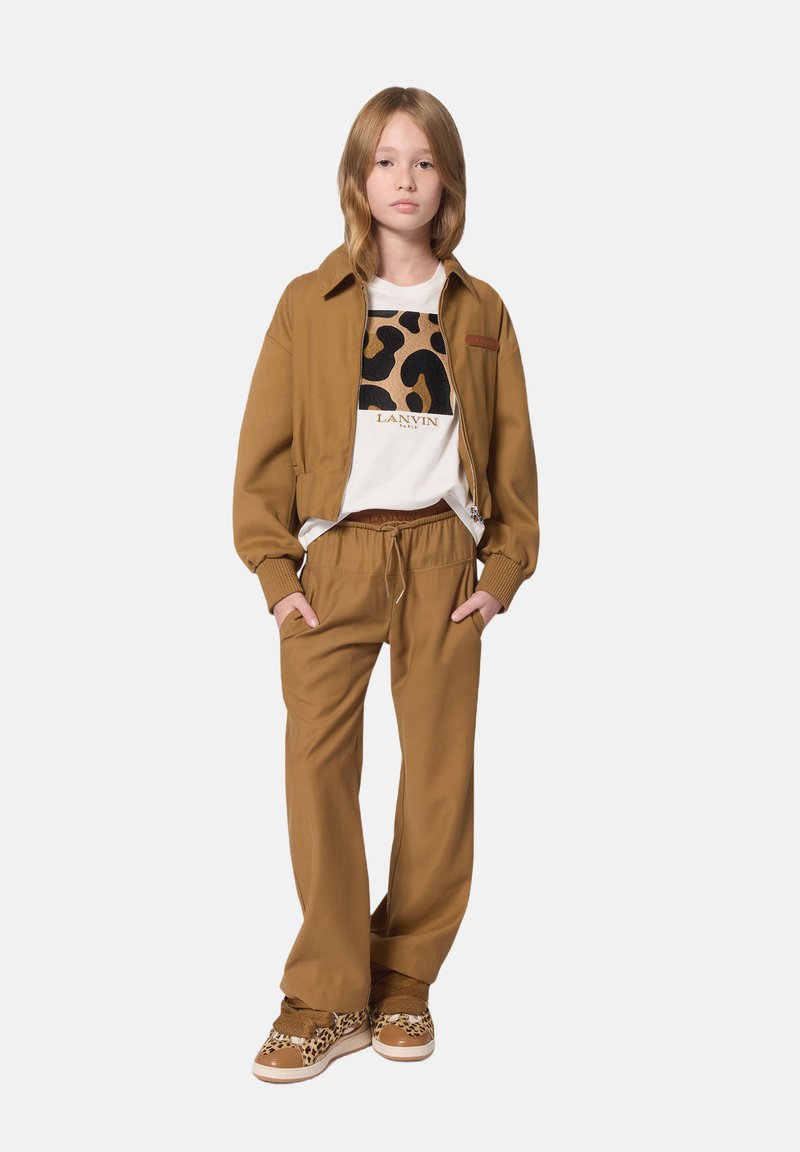 Braune Bomberjacke mit ribbed Bündchen, weißes T-Shirt mit einem Leopardenmuster, passende braune Jogginghose mit Kordelzug und gepunktete Sneakers.