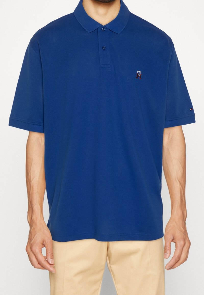 Homme portant un polo bleu à manches courtes avec col et un pantalon beige, debout devant un fond uni.