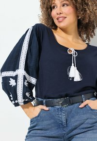 Marinblå topp med texturerade ballongsärmar, vit broderi och tofsdetaljer. Matchad med högmidjade jeans och ett svart bälte.