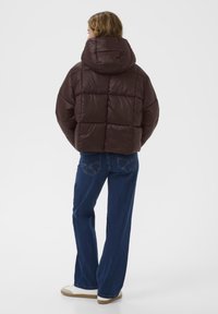 Brūns pufīgs pārklājs ar kapuci, valkāts kopā ar ziliem taisnā griezuma džinsiem un baltām apaviem, skatīts no aizmugures.