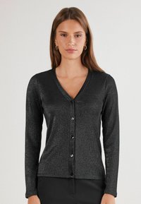 Žena v černém kardiganu s dlouhým rukávem a výstřihem do V, které je zapnuté a má jemný lesk, stojící na pozadí s neutrální světlo.