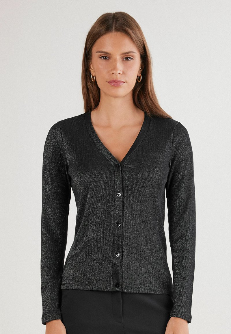 Žena v černém kardiganu s dlouhým rukávem a výstřihem do V, které je zapnuté a má jemný lesk, stojící na pozadí s neutrální světlo.