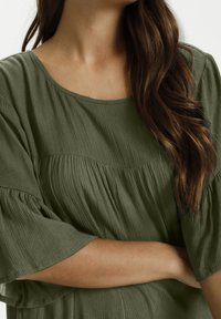 Mujer con cabello largo castaño que lleva una blusa texturizada verde oliva con mangas holgadas, con los brazos cruzados a la altura de la cintura.