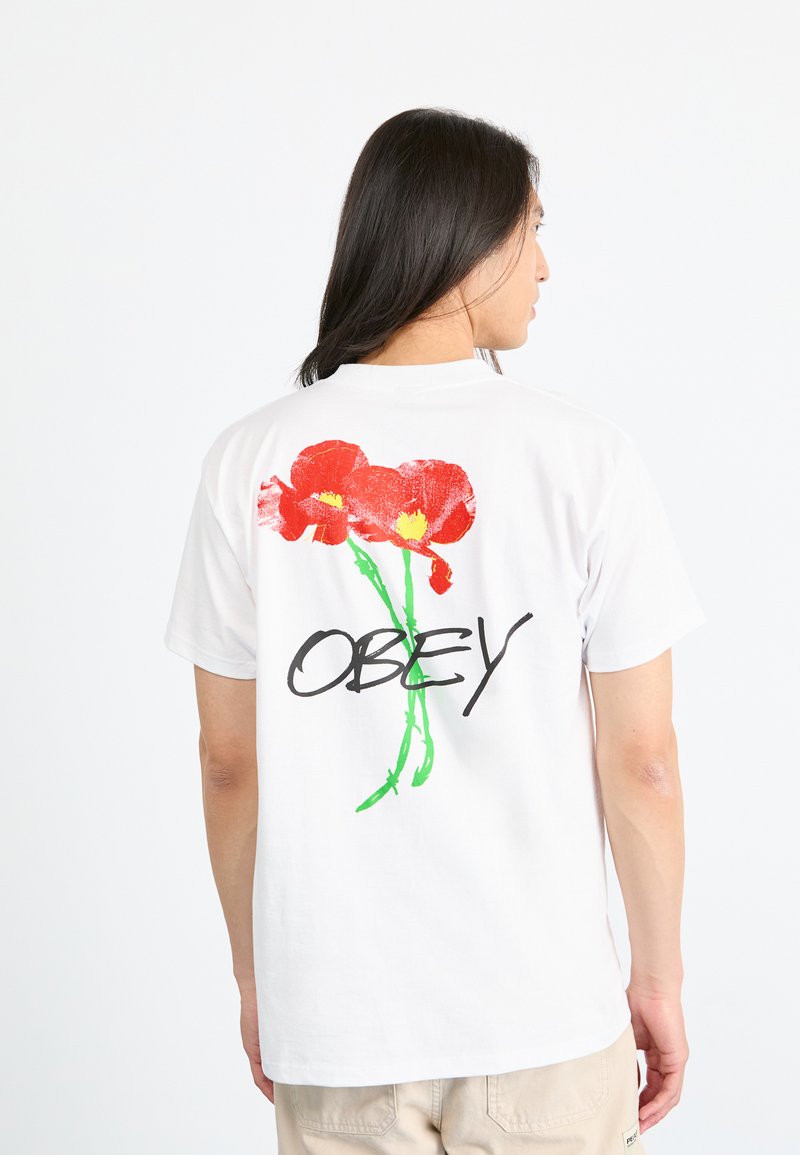 Balta kokvilnas T-kreklā, kura aizmugurē ir spilgta sarkana un dzeltena ziedu grafika, kopā ar vārdu "OBEY" treknā melnā burtnē.